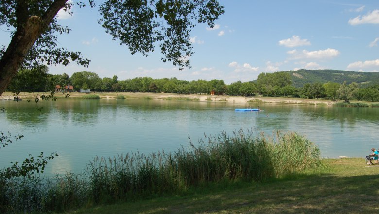 Sea battle, © Gemeinde Langenzersdorf A peaceful lake with trees and a jetty in the background, surrounded by green countryside.