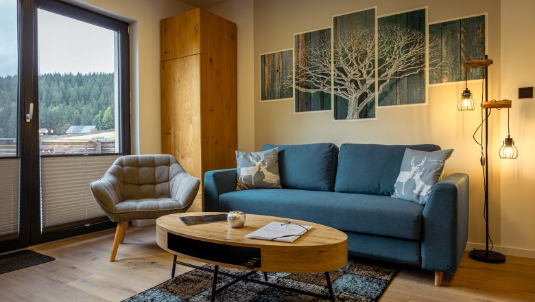 Ötscher Lodge, © Niederösterreich Werbung / Maximilian Pawlikowsky Cozy living room with blue couch, armchair, wooden table and forest view through the window.