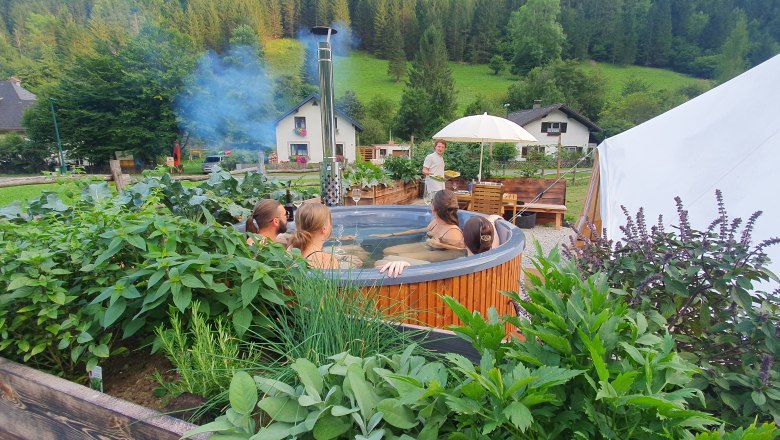 Glamping Wilderness Center Nasswald, © Jonathan Clark People in a round wooden whirlpool surrounded by plants, with a tent and houses in the background.
