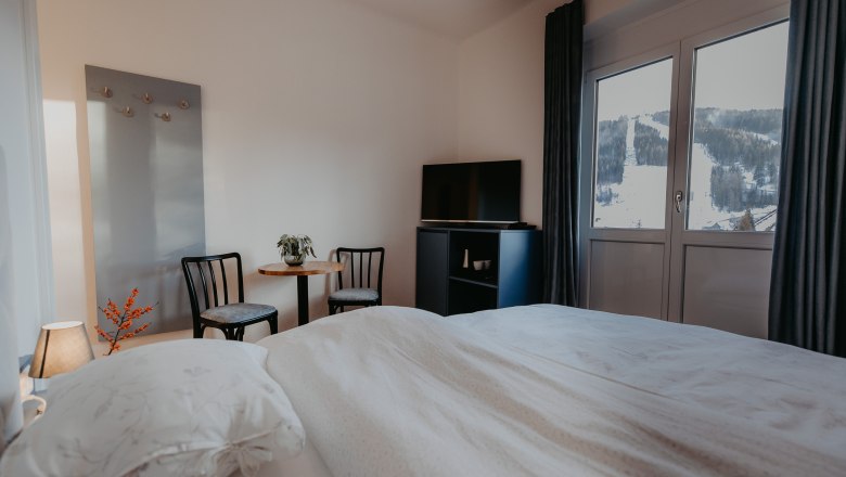 View of the World Cup slope, © Der Löffler - Nicole Seiser A cozy hotel room with a bed, two chairs, a table and a TV. You can see a snow-covered ski slope through the window.