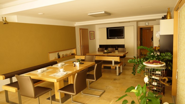 Breakfast room at the Lugerhof, © Lugerhof A breakfast room with wooden tables, chairs and plants, decorated with crockery and a TV on the wall.