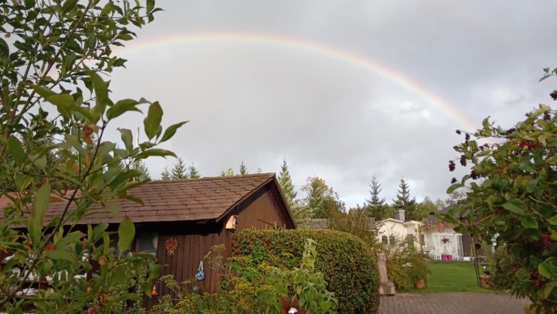 Rainbow over the garden, © Dagmar Scheucher A rainbow stretches over a garden with a wooden house and plants.