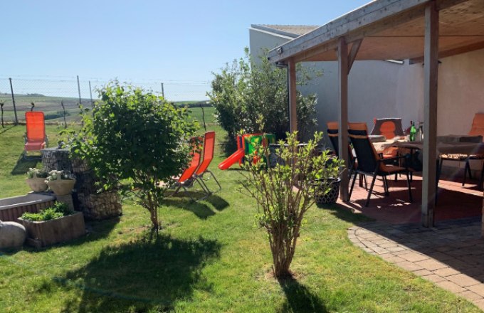 Garden, © Reinhard Gurschka A sunny garden with lawn, shrubs, sun loungers and a covered terrace with table and chairs.