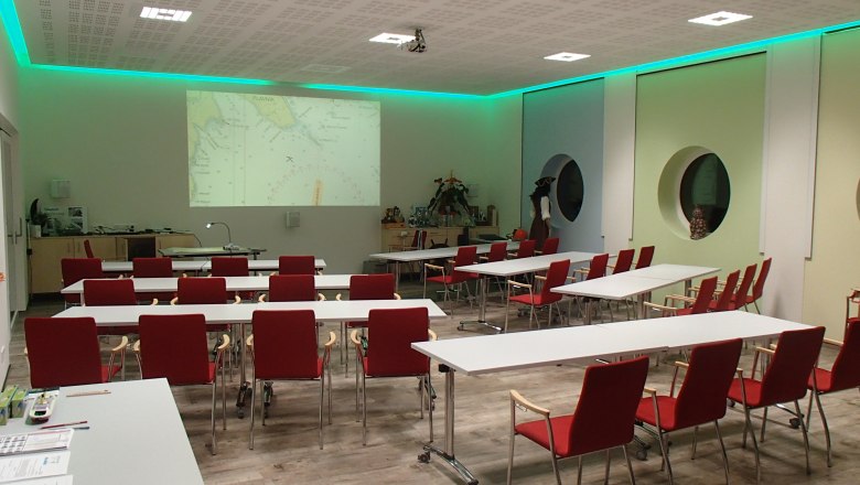 Training room of the Karl Hell Shipmaster School, © Schiffsführerschule Ing. Karl Hell Training room with red chairs and white tables, projector shows nautical chart on the wall.