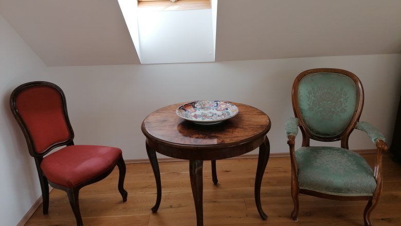 Reading area in the "garden room", © Wiener Alpen Two antique chairs and a round wooden table with a decorative bowl in a room with a skylight.