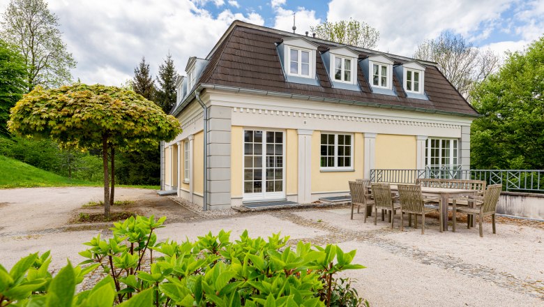 Cavalier houses, © Wiener Alpen / Christian Kremsl Yellow cavalier house with mansard roof, surrounded by trees and garden furniture in the foreground.