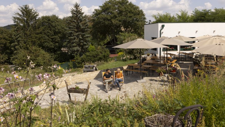 Winter wine tavern, © Weinviertel Tourismus GmbH / Schwarz-König A couple sits on deckchairs in a garden with tables and parasols, surrounded by trees and flowers.
