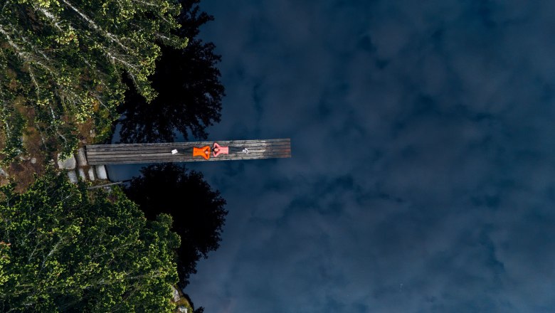 Bull pond, © Waldviertel Tourismus, Melanie Többe Two people are lying on a footbridge over a dark pond, surrounded by trees.