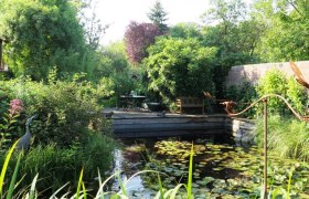 Place by the pond, © Natur im Garten/Martina Liehl-Rainer An idyllic garden with a pond, water lilies and lush vegetation. In the background are a table and chairs on a terrace.