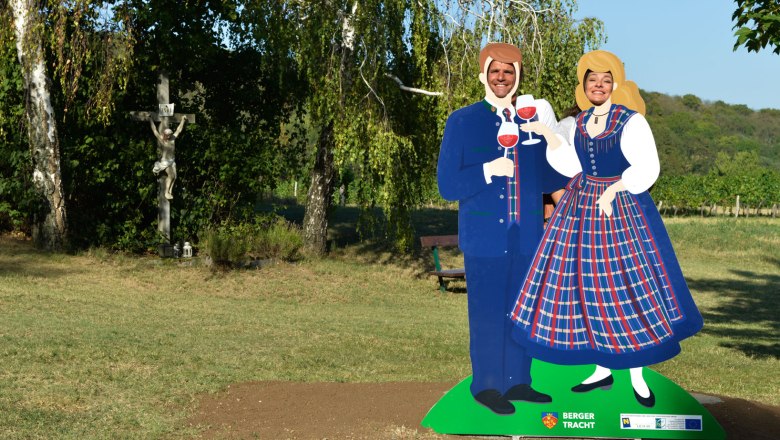 Berger Tracht, VIA.CARNUNTUM., © Donau Niederösterreich, Steve Haider Two people pose behind a cardboard figure in traditional costume with glasses of wine. A crucifix and trees in the background.