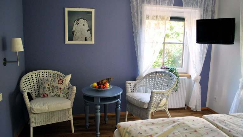 Bed and breakfast Butter Room 1, © Familie Butter Cozy room with blue walls, two wicker armchairs, a round table with a fruit bowl and a window with white curtains.