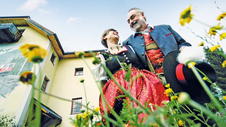 Naturhotel Steinschalerhof, © weinfranz.at A couple in traditional costume stand in front of a yellow building surrounded by flowers, smiling at each other.