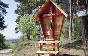 Red Cross Piesting, © MG Markt Piesting/Werner Holy Wooden cross with red beam and carved roof in a forest landscape.