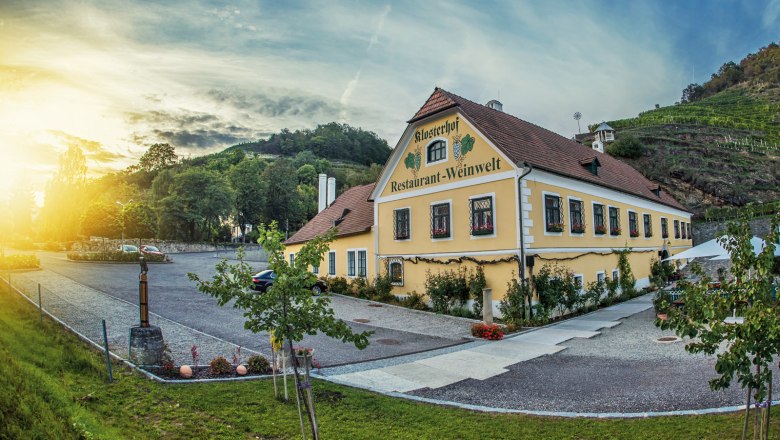 Exterior view, © Klosterhof Exterior view of a yellow building with the inscription 'Klosterhof Restaurant-Weinwelt', surrounded by trees and vineyards at sunset.