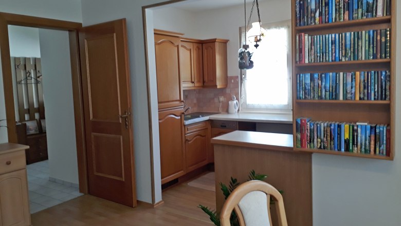 Kitchen, © Barbara Muck Interior view of a kitchen with wooden cupboards, a bookshelf and a dining table.