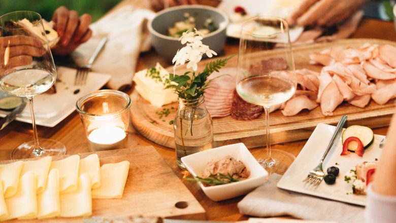 Heurigen snack, © NÖ Werbung / Julius Hirtzberger A table laid with bread, cheese, cold cuts and glasses of wine outside.