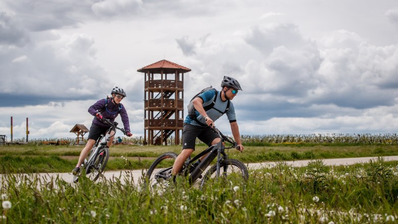 Platt view, © Erwin Haiden Two cyclists ride along a path in front of a lookout tower, surrounded by a green landscape and a cloudy sky.