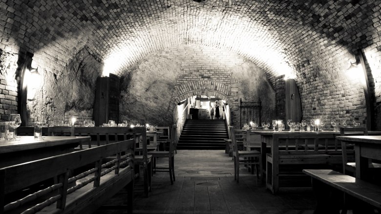 Wine tavern Müllner, © Müllner A rustic wine cellar with wooden tables and benches, illuminated by wall lamps.