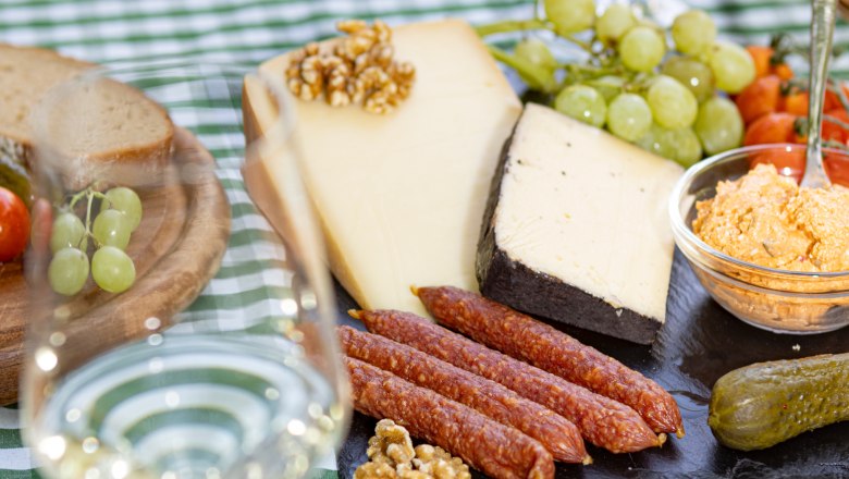 Heurigen snack, © Weinviertel Tourismus GmbH / Schwarz-König A table laid with cheese, sausage, bread, grapes and a glass of white wine on a green and white checkered tablecloth.