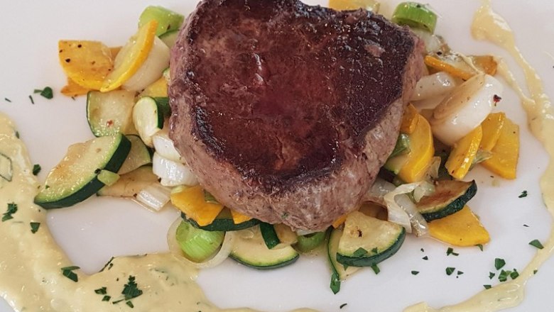 Steak, © Reef & Beef A juicy steak on a bed of roasted vegetables, surrounded by a creamy sauce on a white plate.