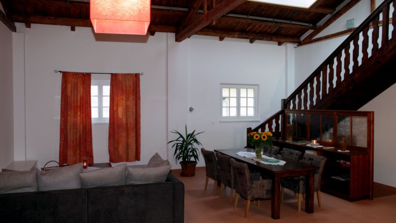 Living and dining area in Loft 1, © Carlos de Mello Living and dining area in a loft with wooden beamed ceiling, red lampshade, gray sofa, dining table with chairs and staircase.