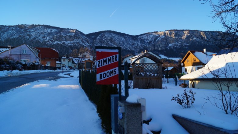 View in front of the house, © Reep Winter landscape with snow-covered houses and mountains in the background. A sign with the inscription 'Zimmer Rooms' is visible.