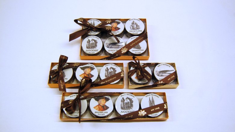 sweet souvenirs, © Konditorei Köller Three golden boxes with round, printed sweets, decorated with brown bows.