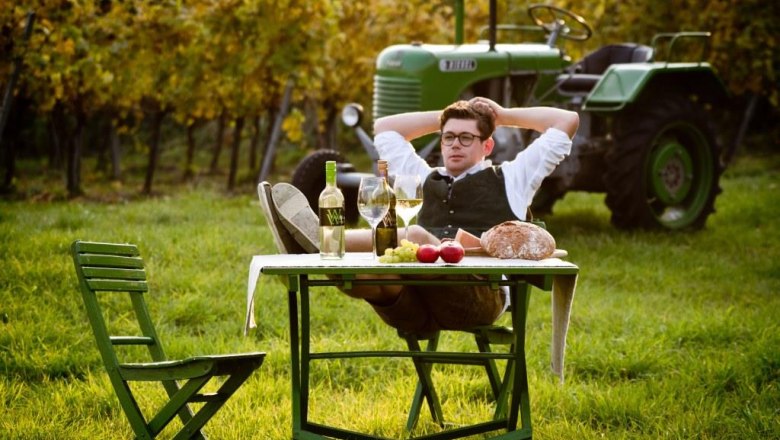 Viticulture Wölflinger, © Fotohaus Hans Krist A man relaxes at a table in the vineyard with wine, bread and apples. In the background is a green tractor.