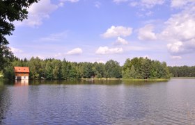 Mitterteich, © Marktgemeinde Hohneich A lake with a house on the shore, surrounded by forest and a blue sky with clouds.