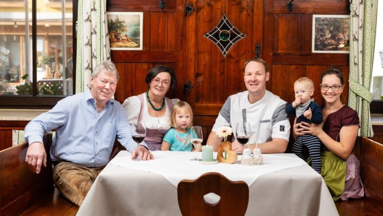 Intergenerational hospitality, © Rita Newman Family sitting at a table in front of a rustic wooden wall. There are 3 wine glasses and decorations on the table.