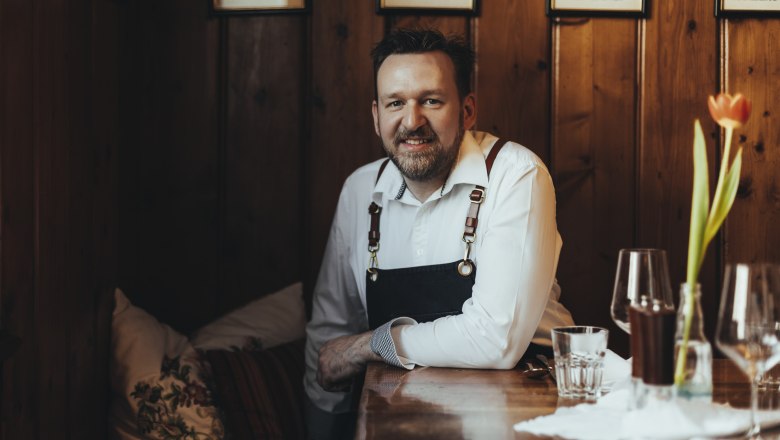 Landlord Christoph Hönig, © Niederösterreich Werbung/David Schreiber A man in a restaurant is sitting at a wooden table, wearing a white shirt and an apron. In the foreground is a vase with a tulip.