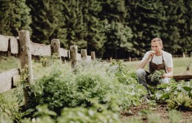 Landlord Markus Kuchner relies on his own fruit & vegetables, © Niederösterreich Werbung/Rita Newman A man in an apron kneels in a vegetable garden and smells herbs, surrounded by green plants and a wooden fence.
