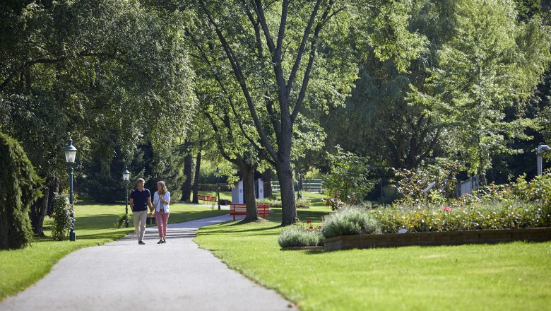 Bad Schönau spa gardens, © Wiener Alpen, Florian Lierzer A couple walks along a path in the spa gardens of Bad Schönau, surrounded by green trees and well-tended flowerbeds.