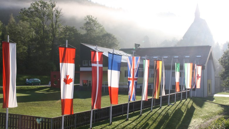 International Wittgenstein Symposium, © Rebecca Gnuechtel, Gemeinde Trattenbach Various national flags in front of a building in a rural setting with fog and trees.