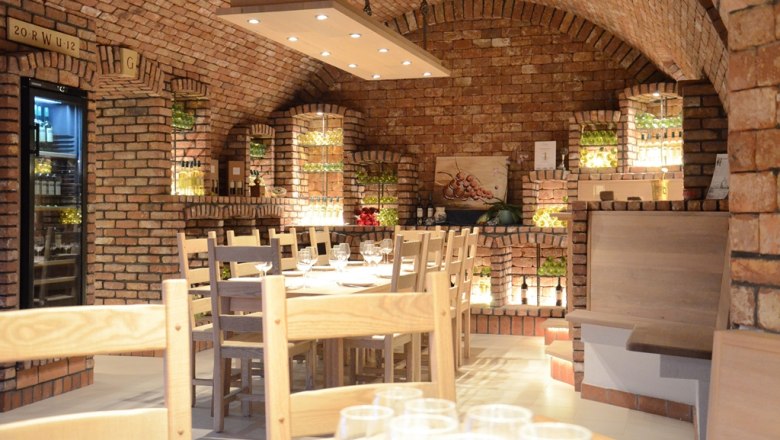 WineLivingRoom, © Weingut Wimberger Cozy wine cellar with brick walls, wooden tables and chairs, illuminated wine rack and decorative elements.
