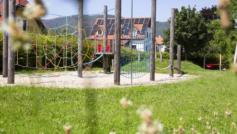 Adventure playground in Joching, © Donau NÖ Tourismus_Barbara Elser An adventure playground with climbing nets and wooden posts, surrounded by a green meadow and houses in the background.