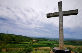 Cone cross, © Weinstraße Weinviertel A large stone cross on a hill overlooking a green landscape and cloudy sky.