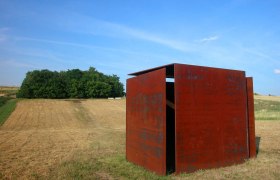 Wind cube house, © Stadtgemeinde Mistelbach A rusty metal sculpture stands in a field in front of a forest under a blue sky.