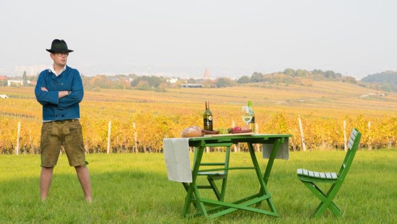 Viticulture Wölflinger, © Fotohaus Hans Krist Man in traditional dress stands on a meadow in front of a table with bottles of wine and bread, vineyards in the background.