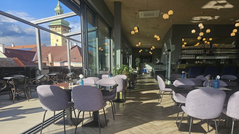 Rooftop indoor area, © Rebecca Freitag Interior of a modern café with glass walls, view of the town hall tower, tables and chairs, wooden floor and decorative lighting.