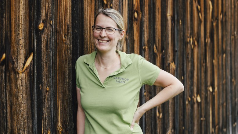 Landlady Irmgard Lang, © Niederösterreich Werbung/David Schreiber A woman in a green polo shirt leans against a wooden wall, smiling.