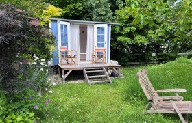 Wild Meadow Show Garden, © "Natur im Garten" A blue garden pavilion with two chairs on a wooden veranda, surrounded by a green meadow and colorful flowers.