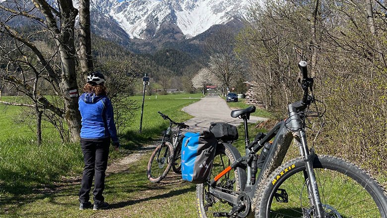 © Angelika Burger A person wearing a helmet stands next to bicycles on a path with snow-covered mountains in the background.