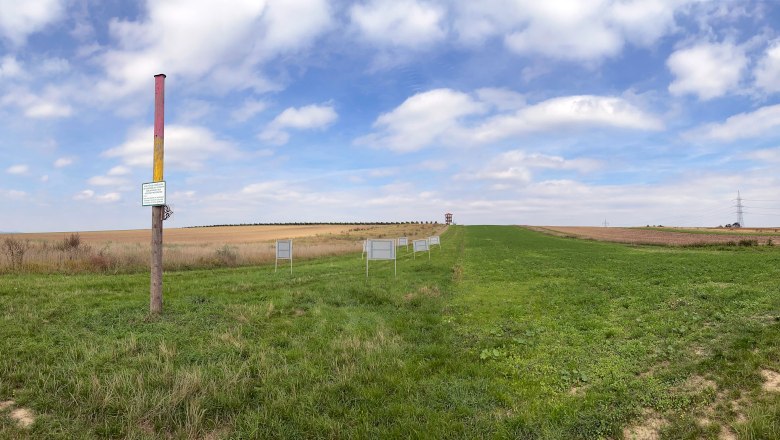 View of the Roseldorf site, © ARDIG Wide landscape with a colorful post and several signs on a meadow under a blue sky.
