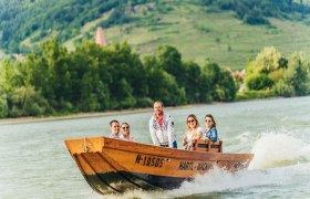 Out and about on the Danube by boat, © Hartl Group of people on a wooden boat on the Danube with a green landscape in the background.