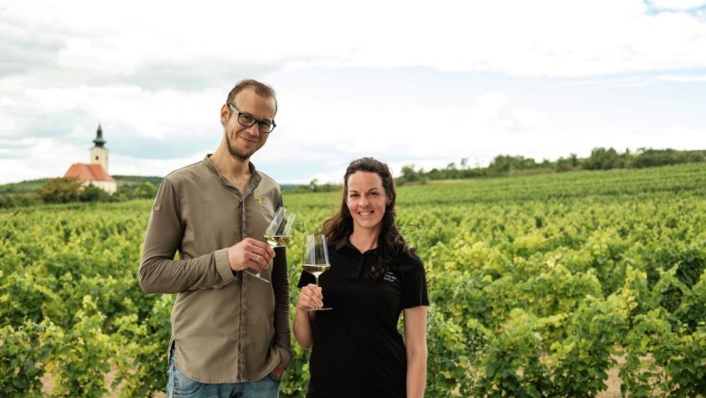 Natalie and David look forward to your visit, © Michael Reidinger Two people stand with wine glasses in a vineyard, with a church in the background.
