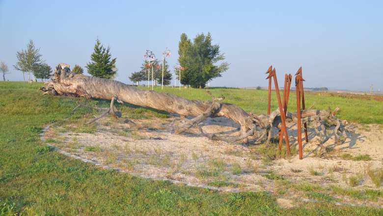 Veltlinerland tree circle, © StadtGemeinde Mistelbach / Mag. Mark Schönmann A large, fallen tree trunk lies on a meadow, surrounded by metal sculptures and trees in the background.