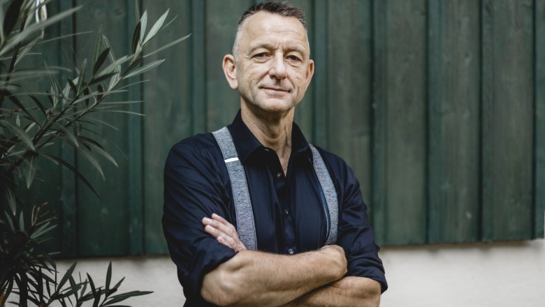 There for the guests: landlord Hans-Joachim Beringer, © Niederösterreich Werbung/David Schreiber A man in a dark shirt and suspenders stands with his arms folded in front of a green wooden wall.