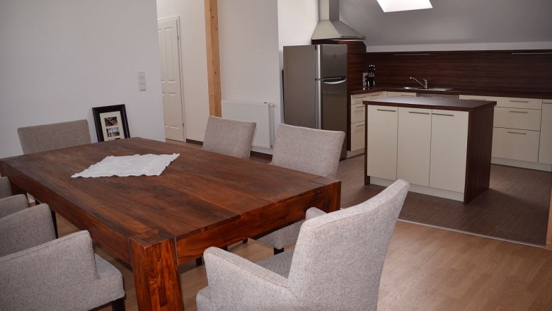 Dining table, © Familie Herrmann Modern dining area with wooden table and gray chairs, adjacent to a kitchen with island and skylight.