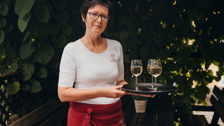 Andrea Ellinger, © Niederösterreich Werbung/Daniela Führer Woman with glasses and tray with two wine glasses outdoors.